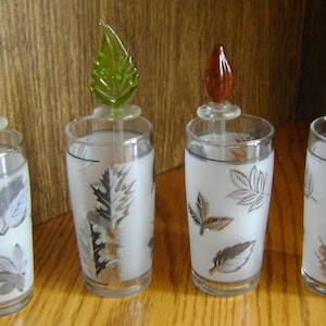 Puede incluir: Cuatro vasos de vidrio esmerilado con diseños de hojas plateadas y tapas de hojas de vidrio de colores. Las tapas son moradas, verdes, naranjas y transparentes.