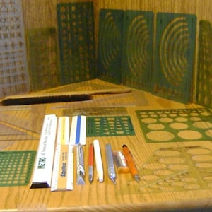 May include: A collection of drafting tools including various green and clear plastic templates with geometric shapes, circles, and ellipses. The set features several rulers, a black brush, a white Metro scale, a Staedtler ruler, and multiple metal craft knives.