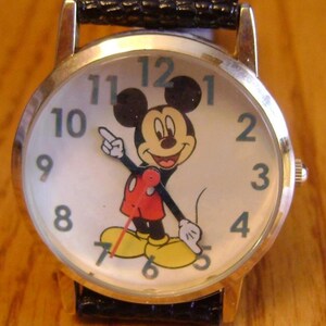May include: A gold-toned wristwatch with a white face featuring a smiling Mickey Mouse character. The watch has black hands and a black leather band. The numbers 1 through 12 are displayed around the face.