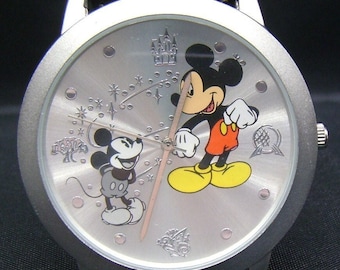 Könnte beinhalten: Silberfarbene Armbanduhr mit Mickey Mouse. Das Zifferblatt hat ein gebürstetes Metall-Finish mit geprägten Disney-Symbolen und zwei Mickey Mouse-Illustrationen. Die Uhr hat ein schwarzes Lederarmband und eine silberne Lünette.