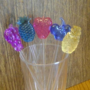 May include: A selection of fruit-shaped cocktail stirrers in a clear glass. The stirrers feature a purple grape cluster, a blue pineapple, a pink strawberry, blue cherries, and a yellow pineapple.