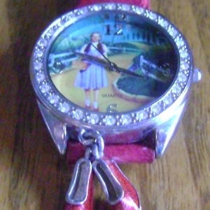 May include: A silver quartz watch with a red leather strap. The watch face depicts a scene from The Wizard of Oz, featuring Dorothy. The watch bezel is studded with rhinestones, and two red ruby slippers hang from the watch.