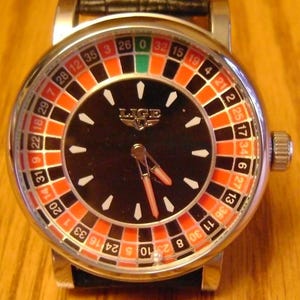 May include: A stainless steel wristwatch with a black face featuring a roulette wheel design. The watch has a silver bezel, white hour markers, and red and black accents. The hands are red, and the watch has a black leather band.