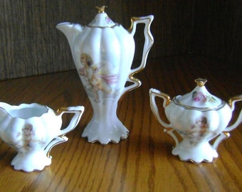 May include: A white porcelain tea set with gold trim, featuring a creamer, sugar bowl, and a tall teapot. Each piece is decorated with cherub illustrations and floral accents, creating an elegant and delicate aesthetic.