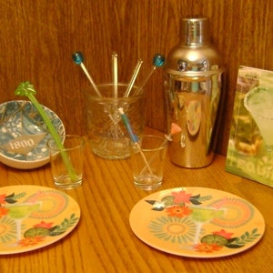 May include: Two colorful plates with a lime wedge and a margarita glass on each. A silver cocktail shaker, two shot glasses, and a glass jar with four blue and green cocktail stirrers are on a wooden surface. A book with the word "Tequila" on the cover is on the surface.