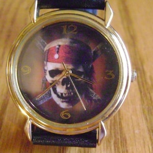 Könnte beinhalten: Eine goldfarbene Armbanduhr mit einem schwarzen Band. Das Zifferblatt der Uhr zeigt ein Totenkopf- und Knochenkreuz-Design mit einem roten Bandana. Die Zahlen 12, 9, 3 und 6 sind auf dem Zifferblatt der Uhr sichtbar.