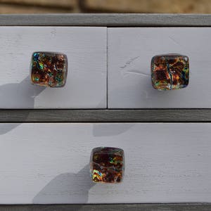 以下が含まれることがあります： 3つの引き出しが付いた小さな灰色の木製キャビネット。各引き出しには、茶色、金色、青色が混ざった正方形の虹色のガラスノブが付いています。キャビネットのフレームは濃い灰色で、引き出しは白く塗装されています。