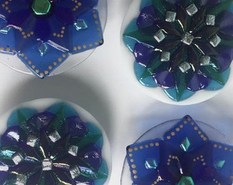 May include: Four decorative glass coasters with blue, green, and silver geometric designs. Each coaster features a starburst pattern with a central jewel-like accent.
