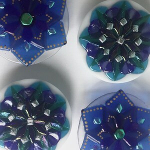 May include: Four decorative glass coasters with blue, green, and silver geometric designs. Each coaster features a starburst pattern with a central jewel-like accent.