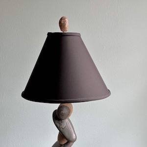 May include: A distinctive table lamp with a black lampshade and a base made from stacked stones. The lamp has a dark brown lampshade and a small stone finial. The base is made of smooth, grey and brown stones.