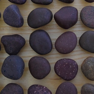 May include: Twenty-five smooth, dark purple and brown stones arranged in five rows of five on a light wood surface. The stones are numbered 1-25.