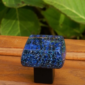 Könnte beinhalten: Quadratischer blauer Glas-Schrankknopf mit schwarzem Sockel. Das Glas hat ein gesprenkeltes Muster und Luftblasen.