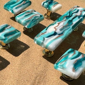 May include: Set of eight teal and white glass cabinet knobs with gold accents. The knobs are in various shapes and sizes, including square and rectangular.
