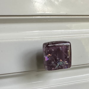 May include: A square-shaped glass cabinet knob with a purple iridescent finish. The knob has a speckled pattern and is mounted on a white cabinet door.