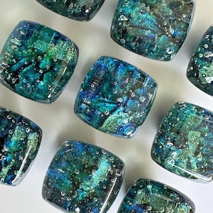 Könnte beinhalten: Eine Sammlung quadratischer Glaskabochons mit einer lebendigen Mischung aus tiefblauen, smaragdgrünen und metallisch türkisfarbenen Tönen. Jedes Stück hat eine glatte, abgerundete Oberfläche mit eingeschlossenen Luftblasen und Glanz.