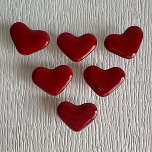 May include: Six glossy, heart-shaped decorative objects in a vibrant red hue are arranged on a textured white surface. The hearts are evenly spaced, showcasing their smooth, reflective surfaces. These decorative hearts are ideal for Valentine's Day.