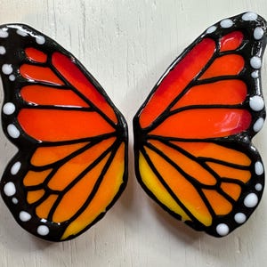 Könnte beinhalten: Ein Paar Schmetterlingsflügel in Orange, Gelb und Rot, mit schwarzen Konturen. Die Flügel haben eine glänzende Oberfläche und weiße Punkte an den Rändern. Das Design erinnert an einen Monarchfalter.