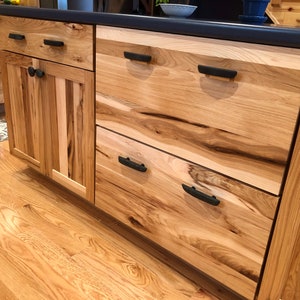May include: Close-up of a kitchen island with wooden cabinets and drawers. The cabinets have a natural wood finish with a knotty grain pattern. The drawers have black metal handles. The island has a black countertop.