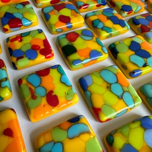 May include: A collection of rectangular glass tiles with a mosaic design. Each tile features a yellow base with a pattern of red, orange, blue, and green shapes. The tiles are arranged in a grid, displaying their vibrant colours.