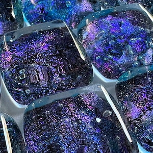 Puede incluir: Primer plano de azulejos de vidrio iridiscente con un esquema de color azul oscuro y morado. Los azulejos tienen una superficie texturizada con pequeñas burbujas de aire atrapadas en su interior.