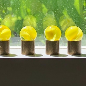 以下が含まれることがあります： 渦巻き模様の入った黄色のガラス玉が4つ、真鍮製の台座に飾られています。玉は白い表面に一列に並んでいます。