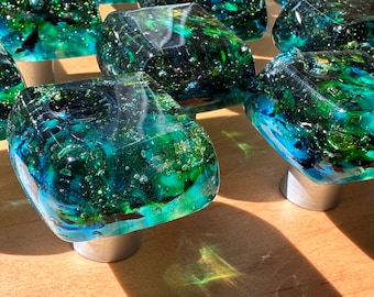 May include: Close-up of six square-shaped glass cabinet knobs with a green and blue marbled pattern and silver bases.