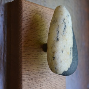 Puede incluir: Un gancho de piedra lisa, blanco y gris, montado sobre una base cuadrada de madera. La piedra tiene una forma natural y orgánica y es una adición única y elegante a cualquier hogar.