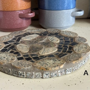 Puede incluir: Un posavasos redondo de piedra gris con un diseño de mosaico de piedras negras y marrones. El posavasos está hecho de pequeñas piezas irregulares de piedra que están dispuestas en un patrón. El posavasos es perfecto para proteger las superficies del calor.