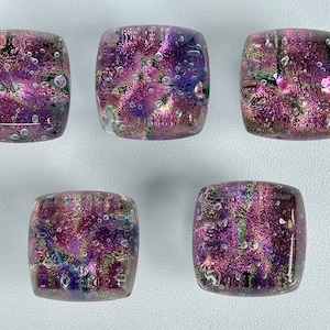 Puede incluir: Cinco azulejos de vidrio cuadrados con un acabado iridiscente morado, rosa y verde. Los azulejos tienen una superficie texturizada con pequeñas burbujas de aire en todo.