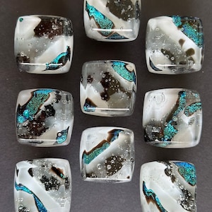 Può includere: Nove piastrelle quadrate di vetro con sfondo bianco e disegni astratti neri e turchesi. Le piastrelle hanno una superficie testurizzata con piccole bolle d'aria.