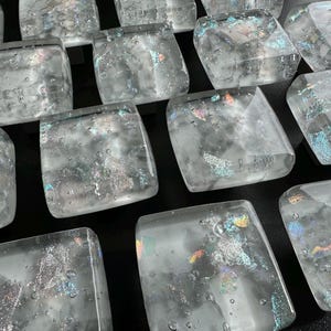 Puede incluir: Una colección de azulejos cuadrados de vidrio con motas iridiscentes y burbujas de aire. Los azulejos son translúcidos y tienen un color gris claro.