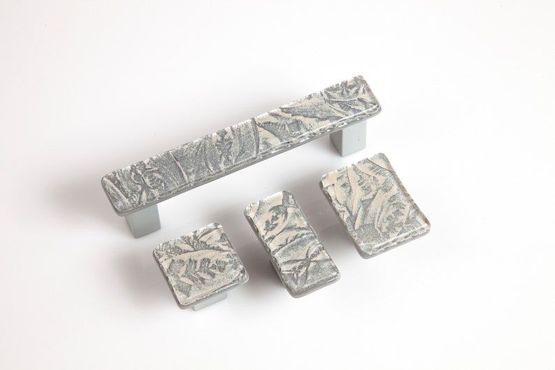 May include: Set of four silver-toned cabinet knobs with a textured, white and gray pattern. The knobs are in various sizes and shapes, including a long rectangular pull.