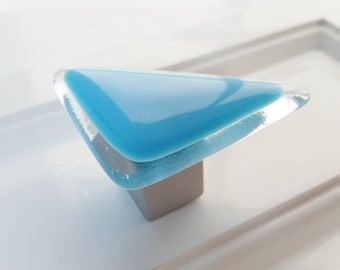 May include: A triangular cabinet knob featuring a smooth, opaque bright blue glass top layer fused to a clear glass base, mounted on a square metallic silver-toned metal post. The design has rounded corners and a polished, reflective surface finish.