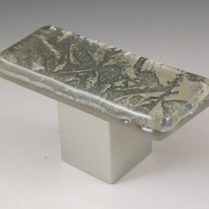 May include: A rectangular glass cabinet knob with a silver base and a textured, light gray glass top. The glass top has a subtle, swirling pattern.