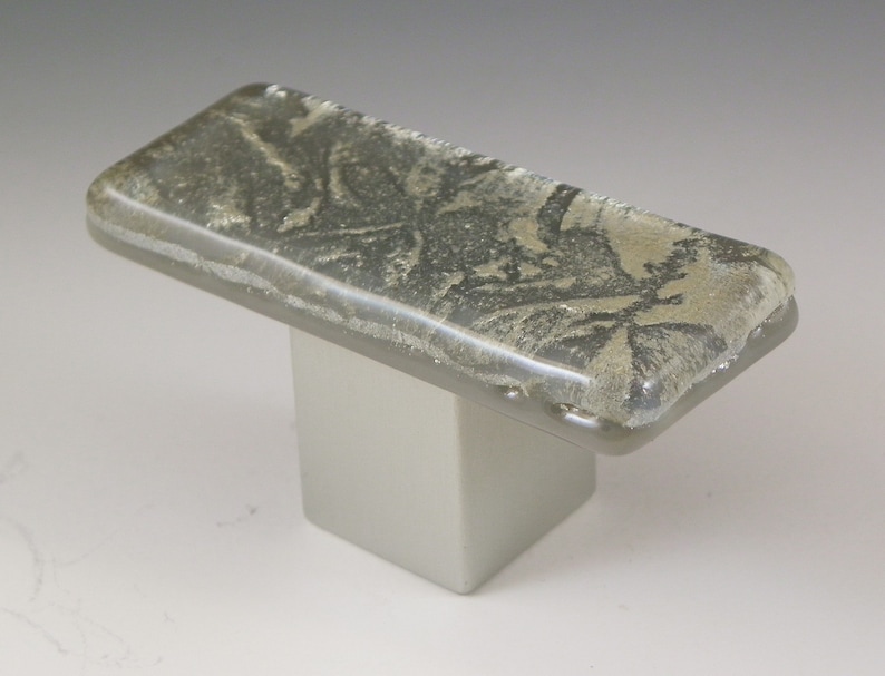 May include: A rectangular glass cabinet knob with a silver base and a textured, light gray glass top. The glass top has a subtle, swirling pattern.