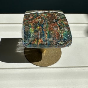 May include: A square glass cabinet knob with iridescent green, blue, and orange colors and small air bubbles throughout. The knob is mounted on a gold metal base.