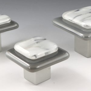 May include: Three cabinet knobs with a grey base and a white marble-like glass top. The knobs are in different sizes and shapes, with one being rectangular and the other two being square.