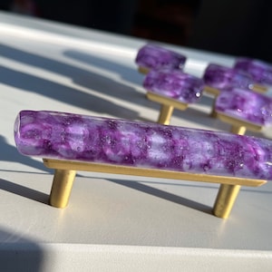 May include: Purple glass cabinet knobs with gold metal hardware. The glass has a swirled, marbled effect.