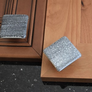 Puede incluir: Dos pomos cuadrados para armario con un acabado gris y blanco moteado. Los pomos son de cerámica y tienen un borde ligeramente elevado.