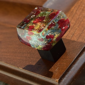 May include: A square glass cabinet knob with red, gold, and blue flecks. The knob is attached to a black metal base.