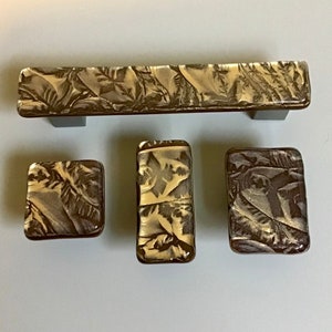 Puede incluir: Conjunto de cuatro pomos de armario de vidrio dorado y marrón con un patrón de hojas. El conjunto incluye una tirador largo y tres pomos más pequeños.
