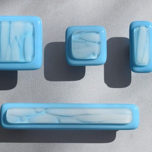 Puede incluir: Conjunto de cuatro pomos de armario de vidrio azul con un diseño de mármol blanco. Los pomos son de diferentes formas y tamaños, incluyendo un cuadrado, un rectángulo y dos círculos.