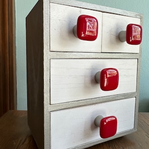 May include: A white wooden dresser with four drawers and red square knobs. The dresser has a silver trim around the edges of the drawers.