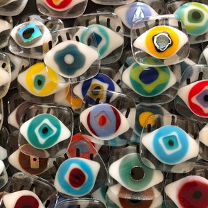 May include: A collection of colorful glass eye charms with white backgrounds and black eyelashes. The eyes are various shapes and sizes and feature different colored centers.
