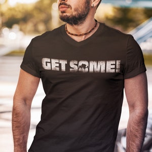 May include: A dark brown V-neck t-shirt with the text "GET SOME!" in distressed white lettering. The shirt features a graphic of a bull. The model is wearing sunglasses and a necklace.