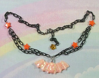 May include: A black chain necklace with orange star accents and a pink glitter bat pendant. The necklace has a layered design and a small gold-colored charm. The background is a pastel rainbow.