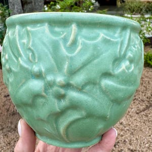 Puede incluir: Una maceta redonda de cerámica verde claro con diseños en relieve de hojas y bayas de acebo. La maceta tiene una superficie ligeramente texturizada y un borde ancho y redondeado. El diseño es de estilo clásico y vintage.
