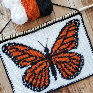May include: A white, orange, and black crochet butterfly wall hanging. The butterfly design is centered on a white background, with orange and black yarn used to create the wings. The piece is mounted on a wooden dowel with a black string for hanging.