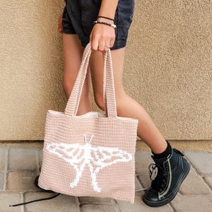 May include: A light pink crocheted tote bag with a white moth design. The bag has two handles and is being held by a person. The bag is made of a textured yarn and has a simple, clean design.