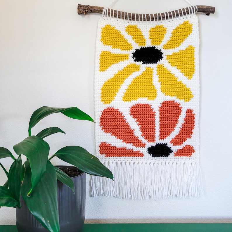 Pu&ograve; includere: Un arazzo da parete all'uncinetto con due grandi fiori stilizzati. Il fiore superiore &egrave; giallo e il fiore inferiore &egrave; arancione. Entrambi i fiori hanno centri neri.